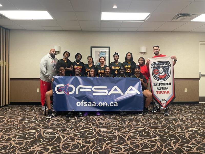 Photo of JCMG girls basketball team posing with team banner and team coaches
