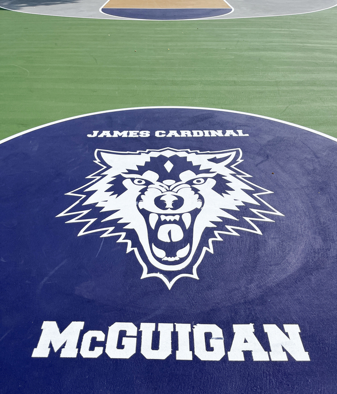 Closeup photo of James Cardinal McGuigan Catholic High School's outdoor basketball court, showing the school name and wolf mascot emblazoned in white on blue flooring.