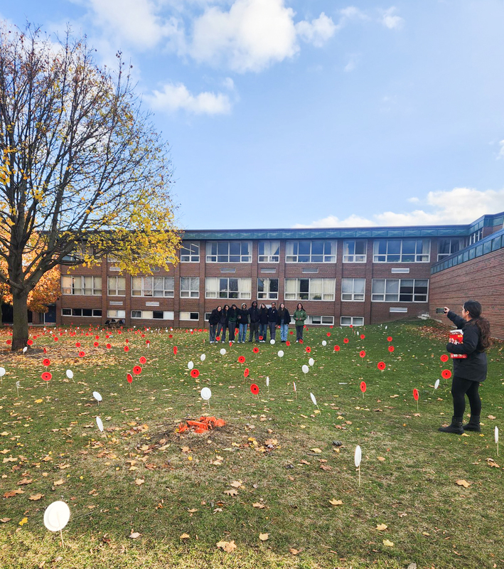 A group of Newman students stand together in the middle of the school grounds where the painted poppies have been planted while a staff member takes a photo of them